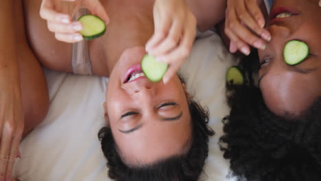 laughing diverse female friends relaxing with cucumber slices during spa day at home