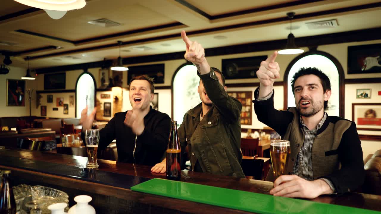 tre tifosi di calcio stanno guardando la partita in un bar sportivo, acclamando e festeggiando i gol con la birra. gli uomini sono molto emotivi, urlano e fanno gesti.