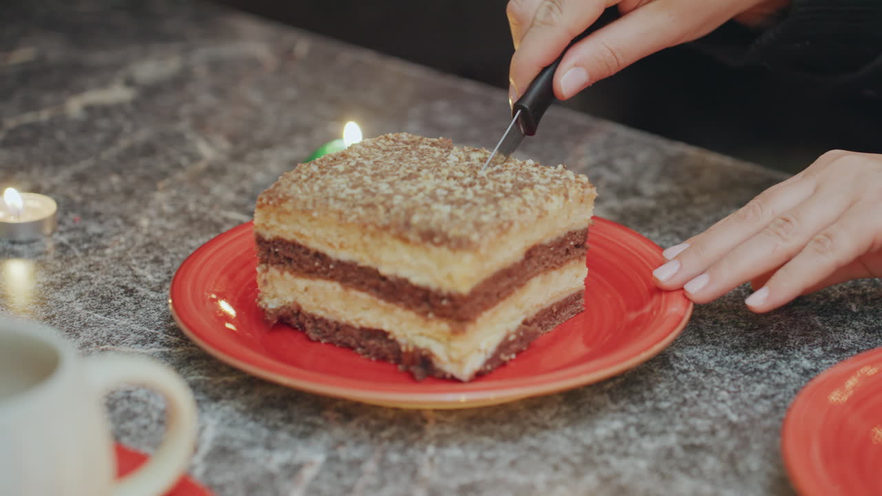 Kırmızı tabakta yakın çekim pasta dilimi. Eller nazikçe dilimi düzeltiyor, yakınında bir kahve kupası, yanan yeşil bir tealight mum. Mermer tezgah yüzeyi, sıcak, yumuşak ışık ve samimi bir sabah fırın atmosferi. Ev yapımı katmanlı pasta.
