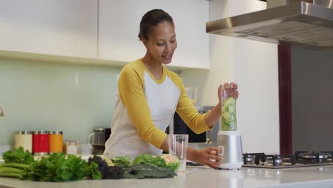 Mixed-race-woman-preparing-healthy-drink,-mixing-fruit-and-vegetables-in-kitchen