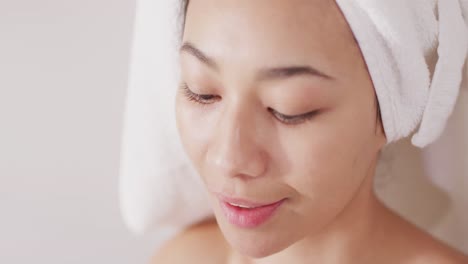 Video-of-portrait-of-smiling-biracial-woman-with-towel-on-hair-applying-face-serum-in-bathroom