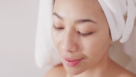 video of portrait of smiling biracial woman with towel on hair applying face serum in bathroom