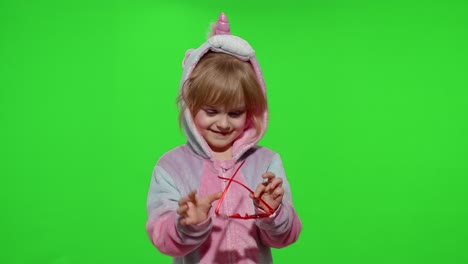 Little-blonde-child-girl-smiling,-puts-on-sunglasses-in-unicorn-pajamas-costume-on-chroma-key