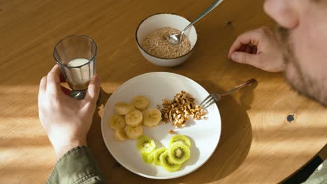 a man is his healthy breakfast with nuts kiwi and bananas on the plate and a glass of milk. filmed from above