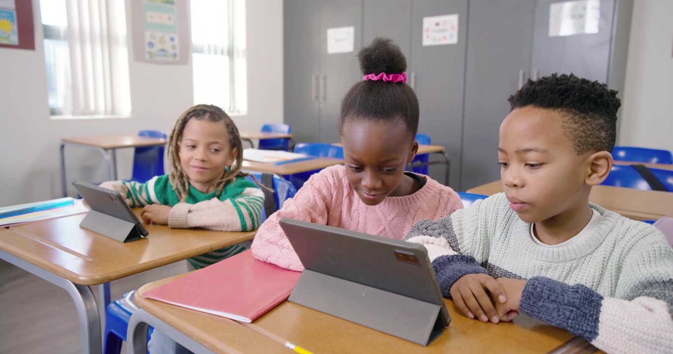 Bambini afroamericani seduti ai banchi, la bambina al centro che guarda il contenuto del tablet, toccando per finire il lavoro