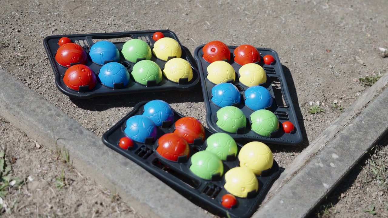 A high-angle view shows three black plastic carrying cases filled with colorful bocce or petanque balls sitting on a sandy outdoor court. The vibrant colorful balls lay organised under bright daylight