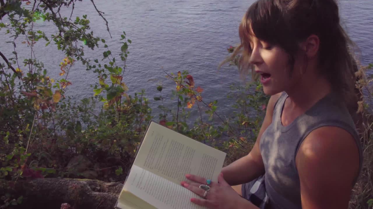 linda chica hipster leyendo un libro en voz alta a la cámara en un parque local junto a un río