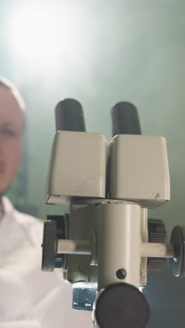 una vista de cerca de un científico examinando intensamente muestras a través de un microscopio en un laboratorio, luego se mueve hacia atrás desde el microscopio, el científico está en foco, con una bata blanca de laboratorio