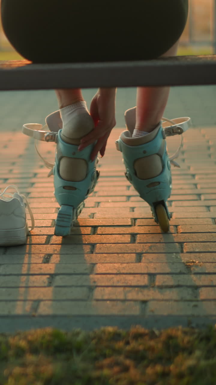 Back view of lady seated on bench in warm evening light removing right roller skate with one hand resting on bench, two white sneakers on ground beside her, sunset glow, and blurred iron railing