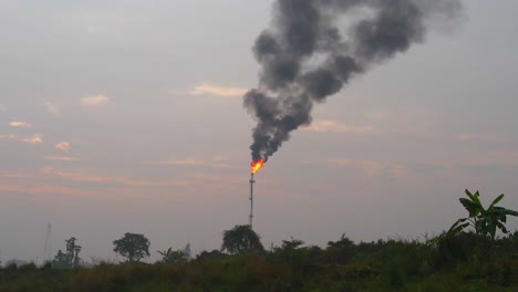 aerial view rising to petrochemical refinery flaming smoking flare stack over misty agricultural farmland at sunrise