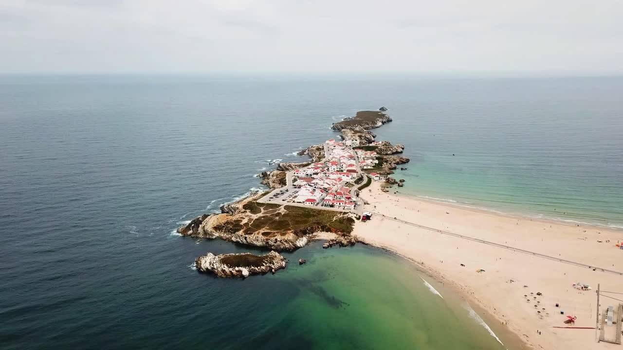 A breathtaking aerial view of Baleal, Portugal, highlighting its scenic coastal peninsula surrounded by clear blue waters, sandy beaches, and a charming village with red-roofed houses
