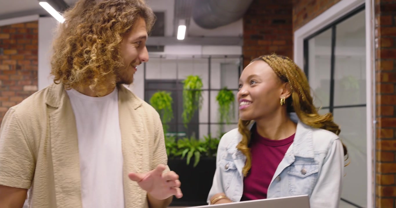 Una donna in giacca di jeans, tra colleghi diversi, mostra un laptop e revisiona i contenuti per un progetto mentre cammina nel corridoio