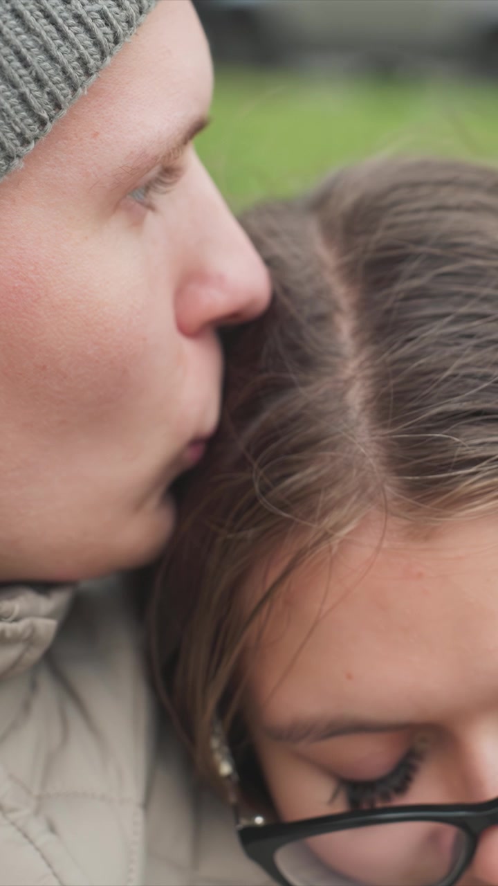 Man supports women who has black framed eyewear. Man wears gray knit hat while woman has brown hair and glasses. Scene takes place possibly near open space or garden area