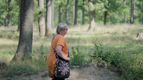 panning shot of an older fat woman with grey hair wearing orange t-shirt and black bag walking through the green forest, holding wooden basket in slow motion during a day