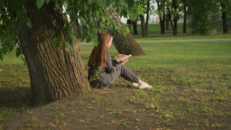 back view of lady seated under tree reading book, hand on page, close to trunk, sunlight softly illuminates her hair and back, legs slightly stretched, surrounded by greenery