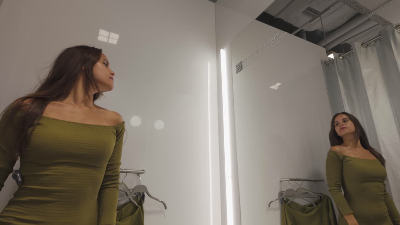A woman examines her outfit in a changing room, adjusting her clothes while looking at her reflection in the mirror.