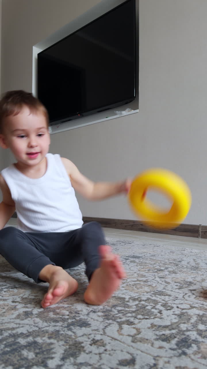 Lovely baby boy in white sleeveless t-shirt and grey pants sits on the floor. Kid puts the toy on his head and drops it. Vertical video.
