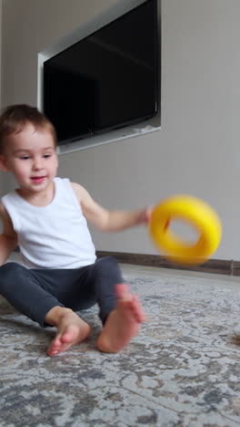 lovely baby boy in white sleeveless t-shirt and grey pants sits on the floor. kid puts the toy on his head and drops it. vertical video.