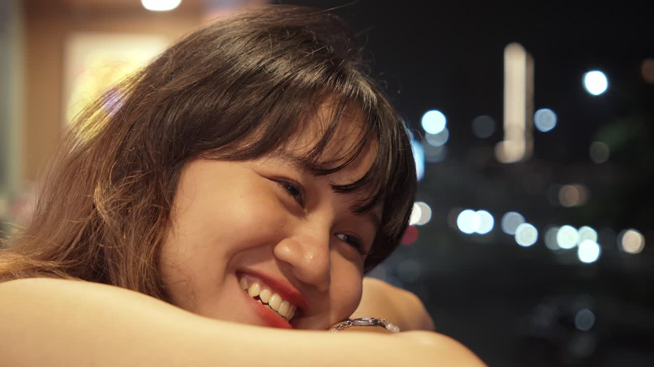 A smiling woman leans on a surface, nighttime bokeh lights sparkle behind her