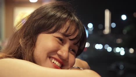 a smiling woman leans on a surface, nighttime bokeh lights sparkle behind her