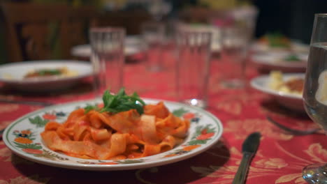 cámara ligeramente panorámica, que muestra una mesa bellamente preparada, un mantel rojo y pasta que se sirve en los platos para una familia numerosa
