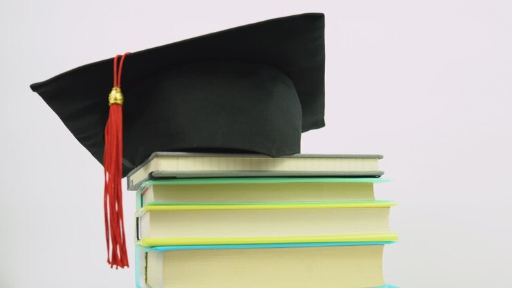 A black square graduate hat with a red tassel lies on a stack of books. Shooting of books, a higher education diploma, a graduate hat and a protective medical mask spinning on a stand