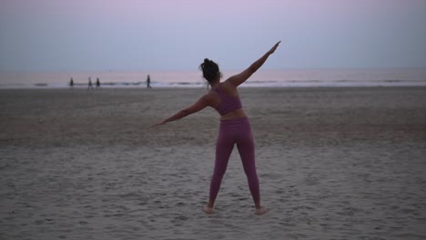 Healthy-woman-in-pink-clothes-doing-yoga,-standing-on-sandy-beach-with-a-beautiful-sea-view