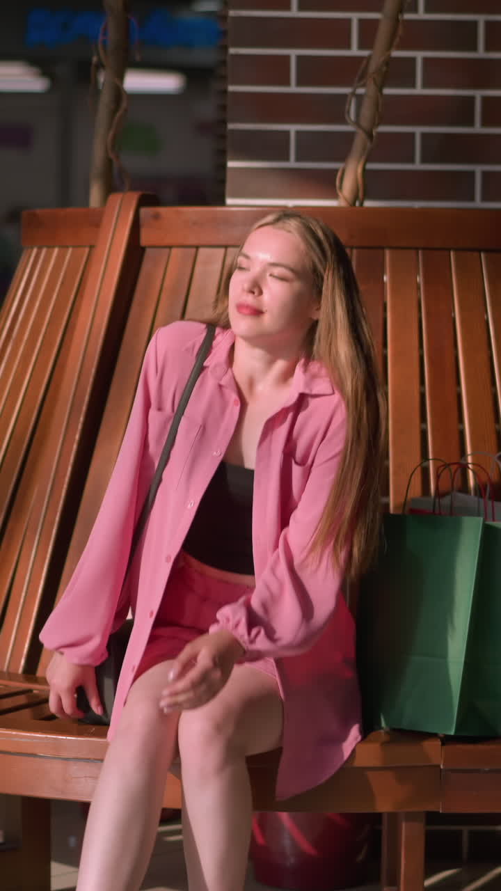 dama vestida de rosa deja caer sus bolsas de compras cerca de un asiento de madera en un centro comercial, ella sostiene un bolso de mano negro se sienta con la mano izquierda descansando en el banco, las piernas ligeramente cruzadas