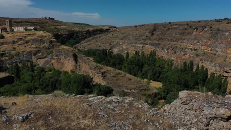 Vuelo-Con-Efecto-Sorpresa-Observando-El-Curso-De-Un-Cañón-Y-Descubriendo-Un-Hermoso-Pueblo-Con-Su-Torre-Y-Sus-Prados-Secos-En-Verano-Con-Un-Cielo-Azul-En-Segovia-España