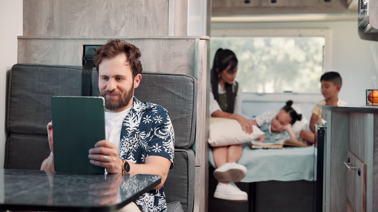 Šťastný muž používající tablet, zatímco rodina odpočívá v obytném voze.