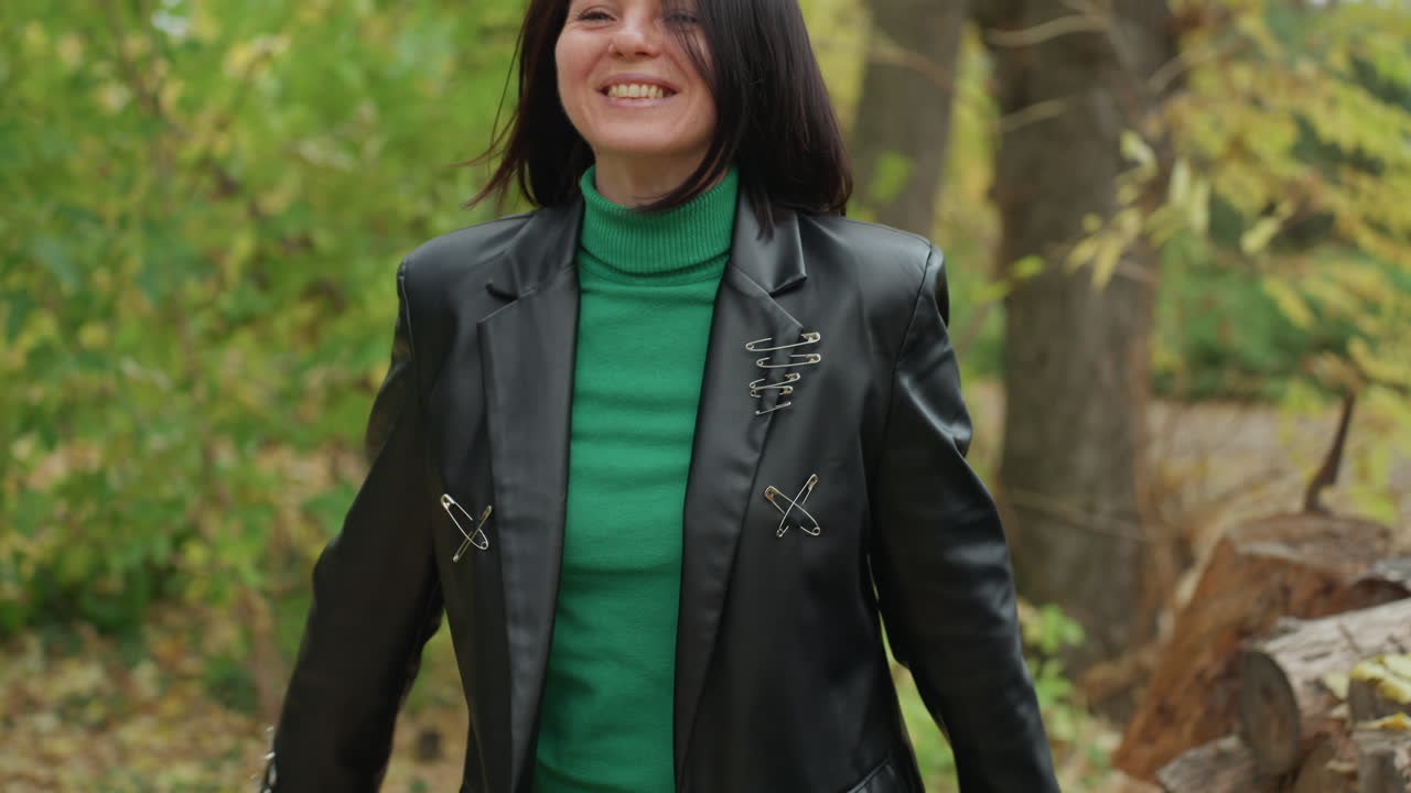 woman in leather jacket walking through autumn forest near stacked woodpile, green sweater and black pants, smiling with confident rocker energy, hands swinging, candid fullbody shot, fallen leaves