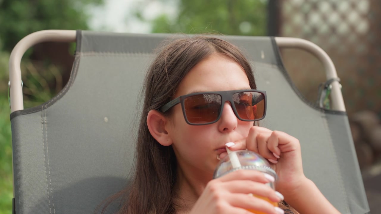 Woman relaxes outdoors on chair reaching for and drinking orange juice from plastic cup with straw. Fresh fruits like cherries and apples are visible in foreground against greenery