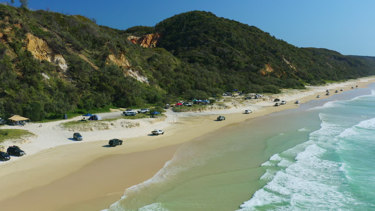 convoy aéreo de autos conduciendo a lo largo de campamentos de playa junto a las olas azules del océano en queensland, 4k drone