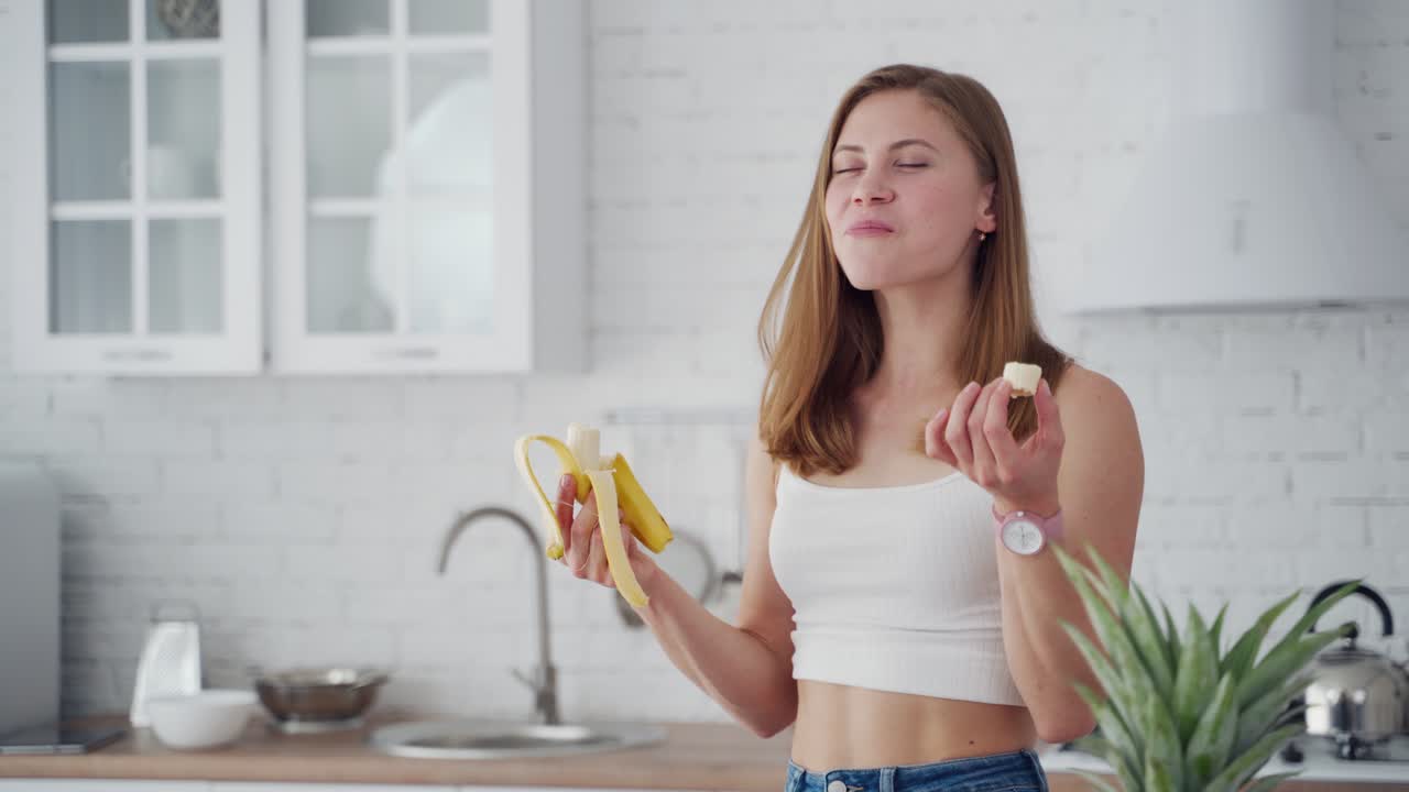 Pretty woman enjoys eating banana. Young female in casual clothing eats fresh fruit in the kitchen. Healthy food concept.