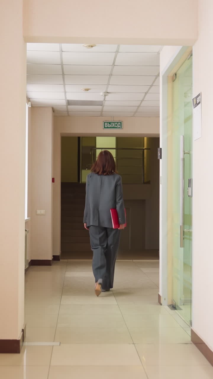 Mulher de negócios de terno cinza caminhando pelo corredor do escritório, segurando uma pasta vermelha, indo para uma reunião ou apresentação com uma postura focada em um espaço de trabalho corporativo moderno durante um dia de trabalho profissional