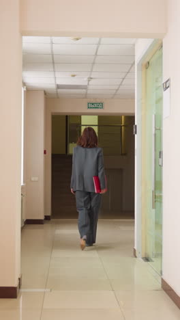 businesswoman in gray suit walking down office hallway holding red folder heading to meeting or presentation with focused posture in modern corporate workspace during professional workday