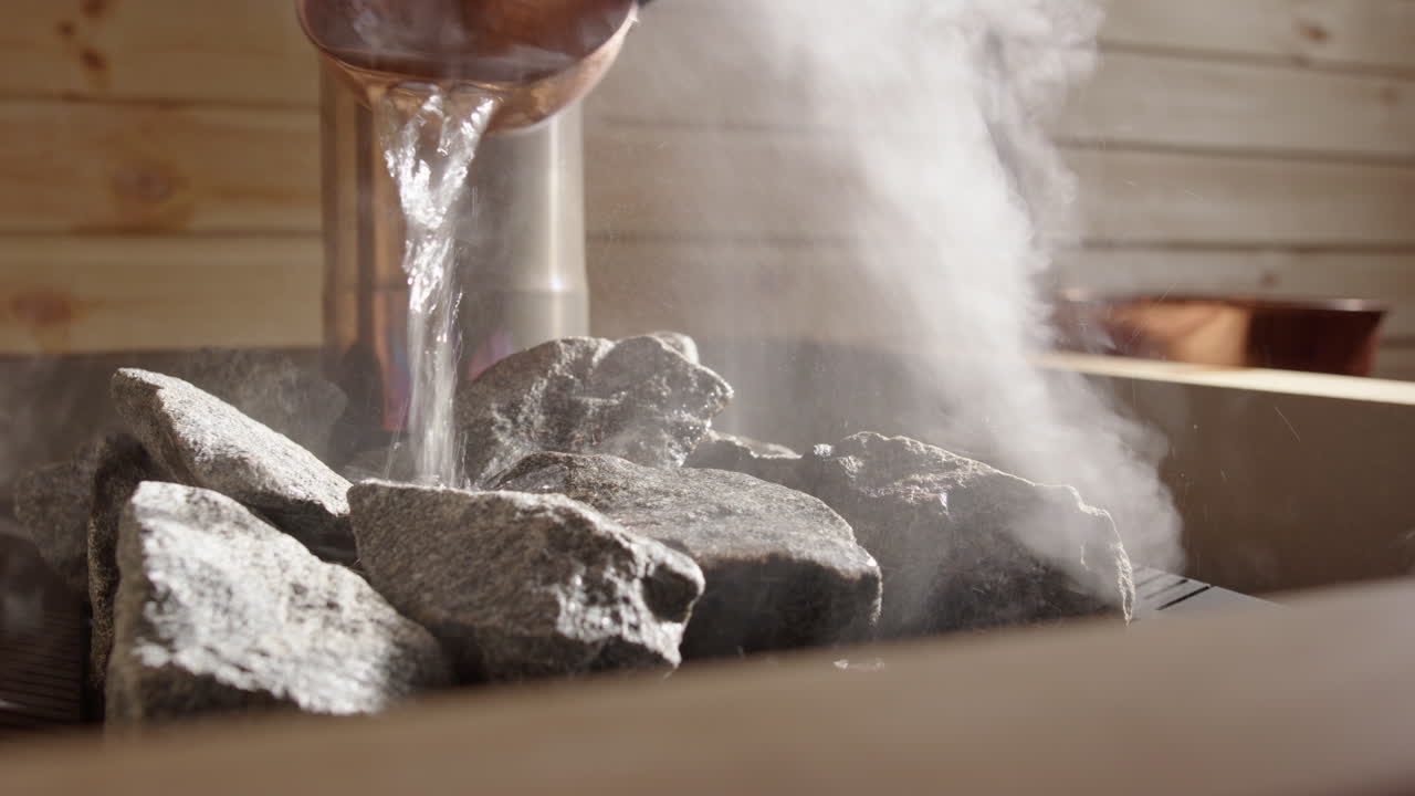 Real time closeup view as copper ladle pours water on hot steaming sauna stones