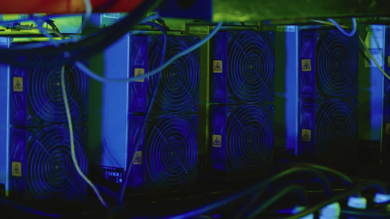 Bitcoin ASIC miners with circular cooling fans and visible power cables lined on a metal shelf, warning stickers attached. Close up shot