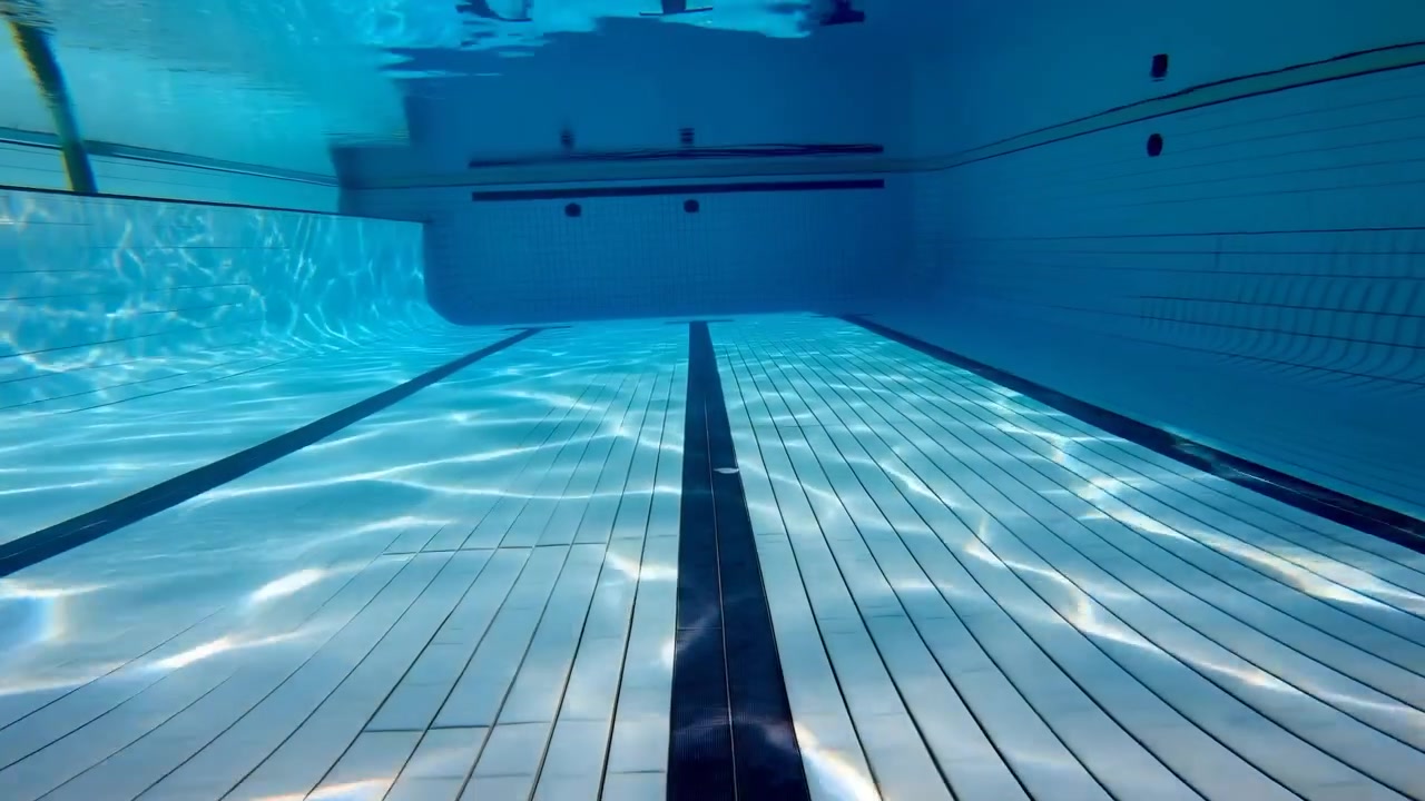 Lav undervandsudsigt i en tom indendørs swimmingpool, der viser en central mørk banemarkering på hvide keramiske fliser, blødt krusende lys, stille blåt vand, ren symmetri og dybde mod bagvæggen.