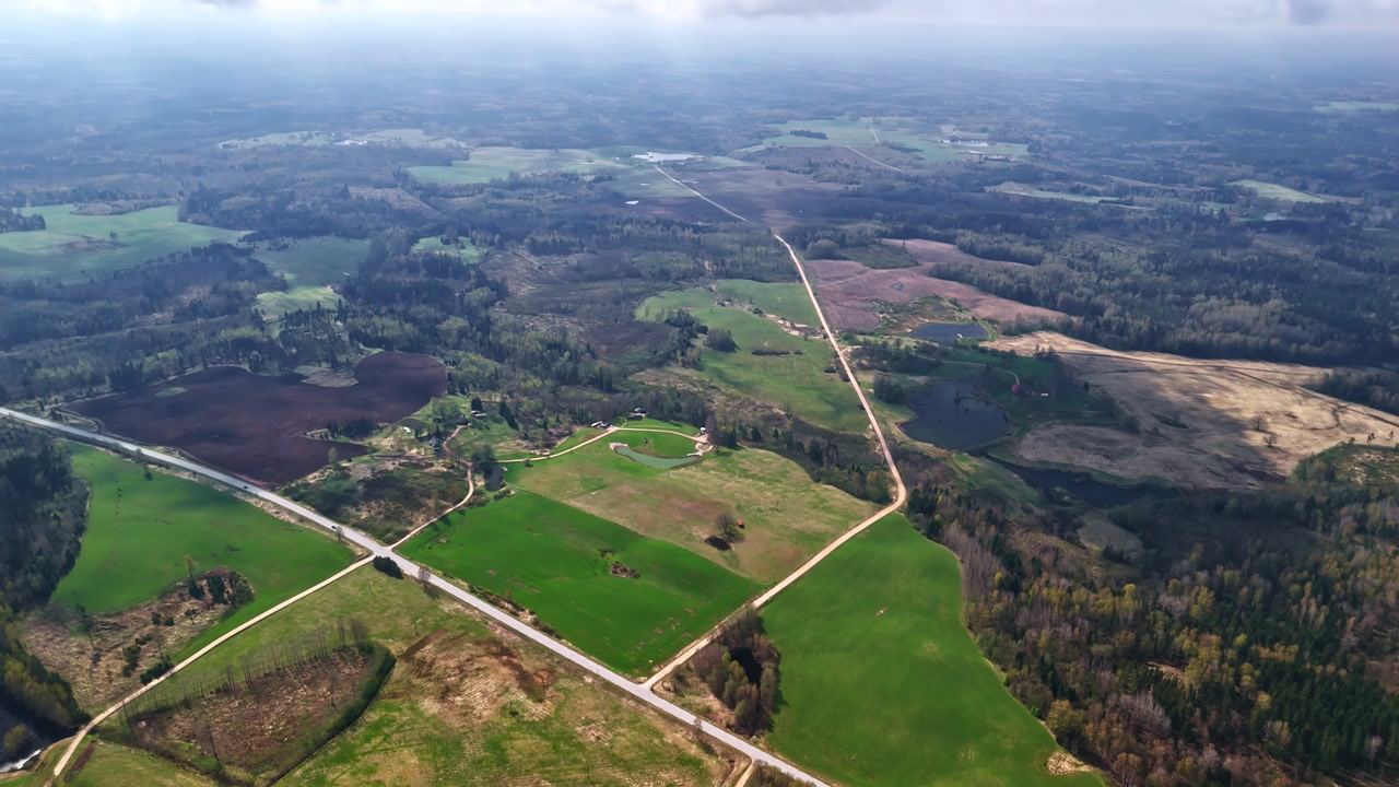 Aerial view of patchwork farmland with crossroads, fields and forest edge under soft daylight. Rural landscape scene with open terrain, winding roads and agricultural land patterns.