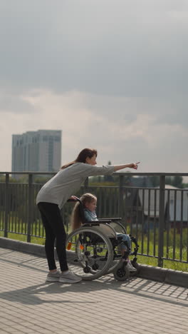 woman shows city architecture to little girl sitting in wheelchair. mother and daughter with cerebral palsy enjoy family trip on sunny spring day