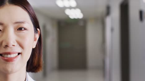 Half-face-video-portrait-of-smiling-asian-female-doctor-in-hospital-corridor,-with-copy-space