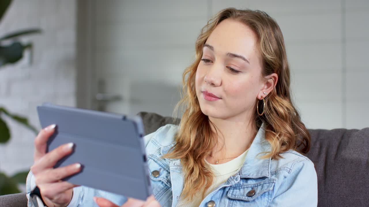 Parikymppinen nainen nostaa tablettia, lukee sohvalla kallistaen sitä samalla kun käsittelee tietoa