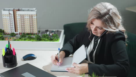 businesswoman multitasking at desk holding phone between ear and shoulder while writing in notebook with polished nails, working in bright modern office with architectural model in background