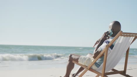 hombre afroamericano senior en una silla de cubierta, usando un teléfono inteligente en la playa, espacio de copia, en cámara lenta