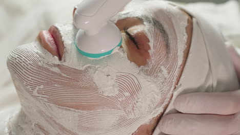 beauty therapist uses vibrating electric facial roller with gloved hand to massage white cream mask on spa client under bright lamp for deep skin rejuvenation and soothing relaxation