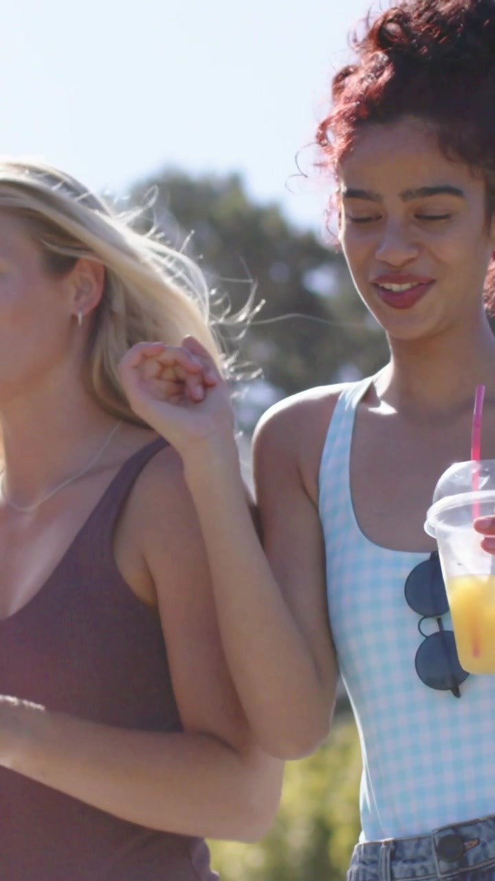 Vertical video: Stepping pool edge, two women walking at pool in white, denim shorts, holding cups