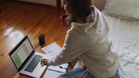 mujer de raza mixta trabajando en casa, sentada en la cama usando una computadora portátil y bebiendo té en la cabaña