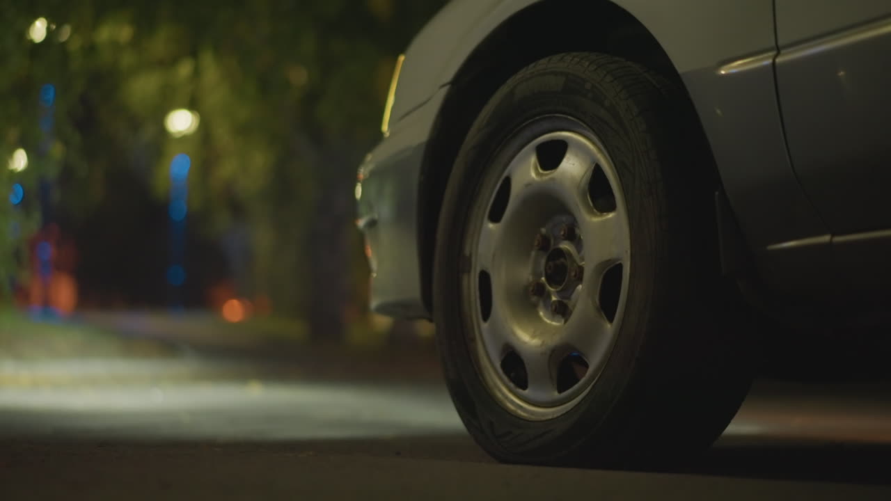 Disparo a neumático bajo en carretera solitaria bajo el brillo de una farola, detalle de la llanta en primer plano con pavimento mojado y borde frondoso, carril vacío que se extiende hacia árboles oscuros, quietud cinematográfica y acciones de encuadre bokeh dispersas