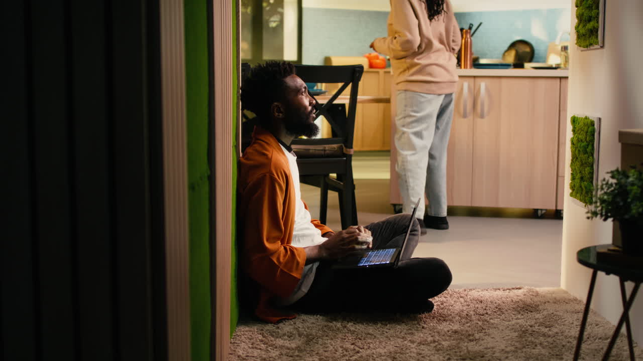 Black young man, sitting on the floor, scrolling on laptop, warm interior